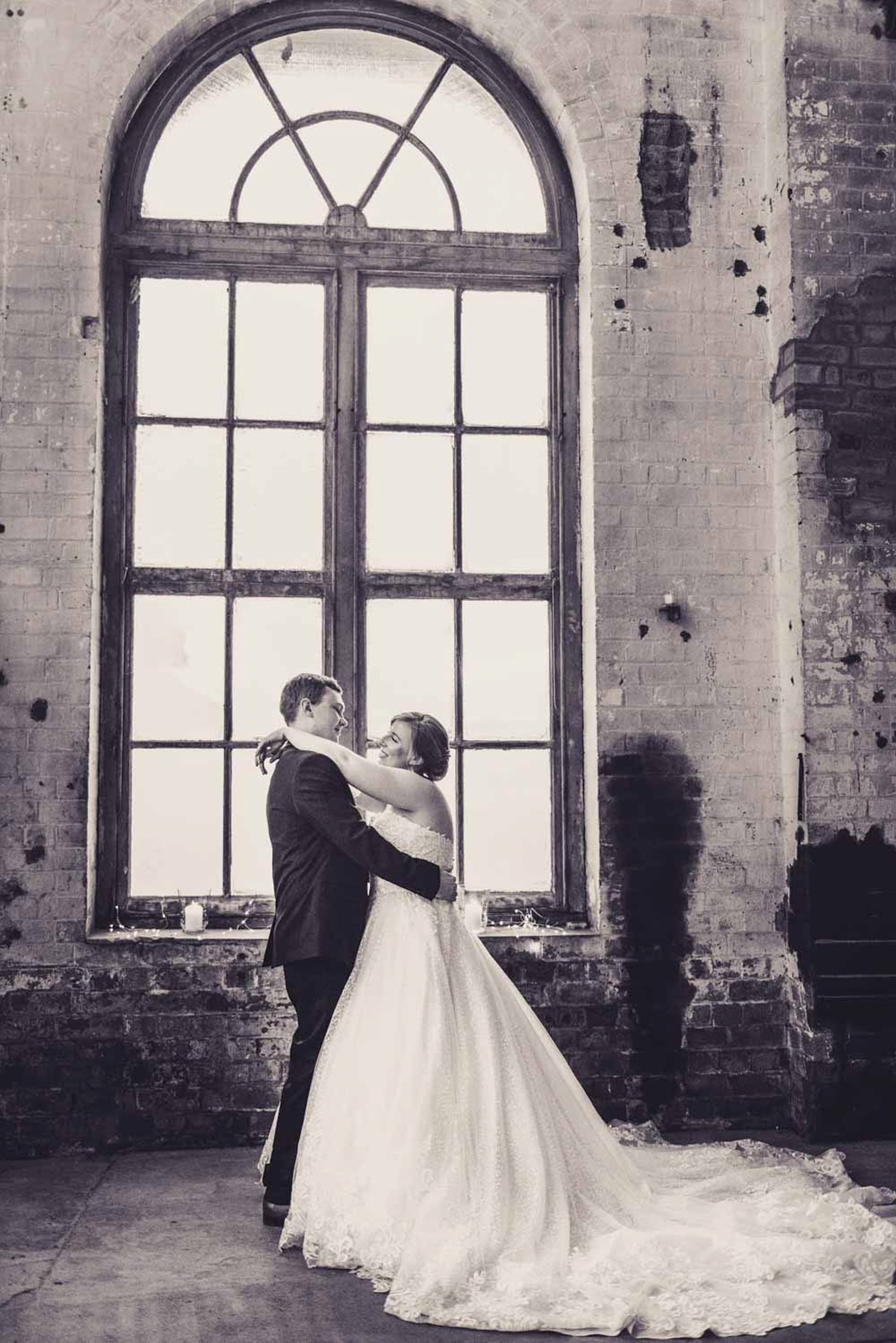 Rhiannon and Lachlan at The Foundations Portland in the Blue Mountains — a real wedding photographed by John Adams, the couple framed by old industrial brick and tall windows in soft afternoon light